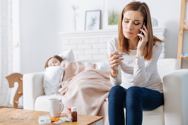 Worried mum on the phone with a child lying down on the sofa behind her